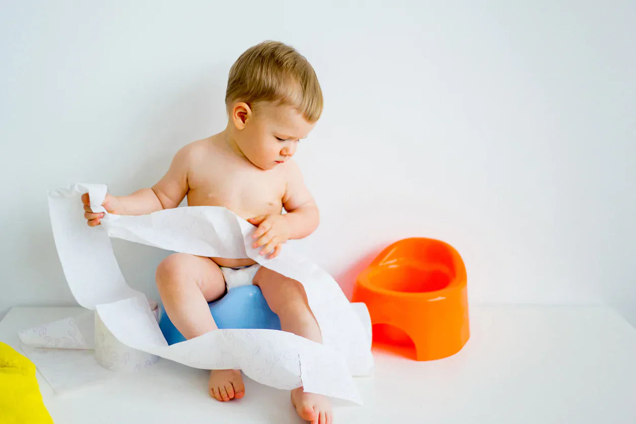 Toddler sitting on a blue potty, playing with toilet paper, while using the Oopsie Heroes daytime toilet training device.