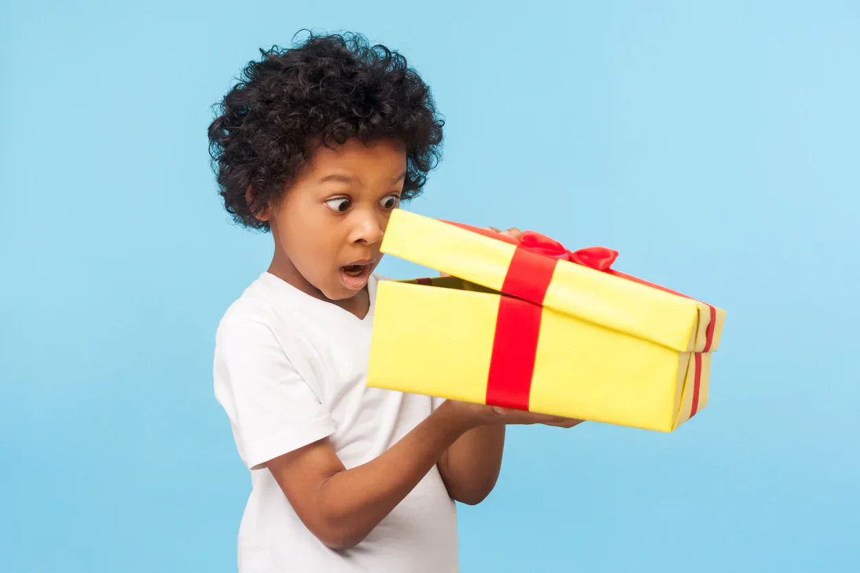 Excited child peeking into a yellow gift box with a red ribbon, symbolising the joy of starting toilet training with a new product.